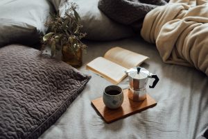 From above of cup of hot coffee with pot on wooden tray near open book and dry plant in cozy bed